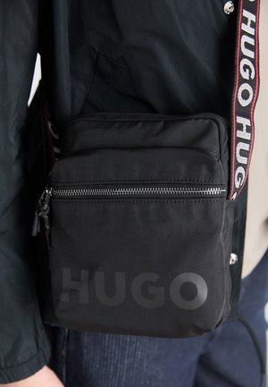 Sac bandoulière noir avec poche avant zippée et texte "HUGO", porté avec une veste noire et une bandoulière de marque affichant "HUGO" en lettres blanches.