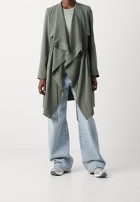 Hellgrüner, drapierter Mantel mit Schalkragen und Kordelzugsaum. Kombiniert mit weit geschnittenen blauen Jeans und grauen Turnschuhen. Glatte Textur.