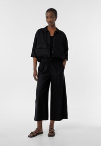 Camicia nera di lino con collo e vestibilità over size, abbinata a pantaloni neri a gamba larga con elastico in vita e tasche.