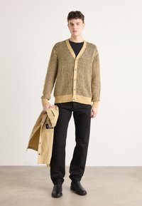 Sweter robiony na drutach w kolorze żółtym z wzorem w kratę, z dekoltem w serek i guzikami. W połączeniu z czarnymi spodniami i beżową kurtką trzymaną w ręku.