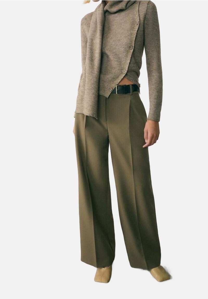 Femme portant une écharpe en maille beige et un pull asymétrique boutonné, associé à un pantalon large vert olive et des chaussures beiges.