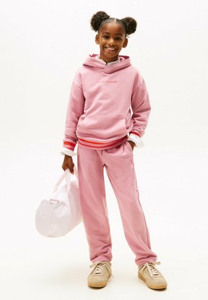 Lichtroze hoodie met het "TOMMY HILFIGER" logo, rood-witte gestreepte boorden. Bijpassende lichtroze sweatpants. Meisje met een witte sporttas.