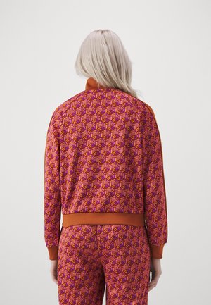 Person med langt lyst hår iført matchende lilla og orange geometrisk mønstret jakke og bukser, stående med ryggen mod, på ensfarvet baggrund.