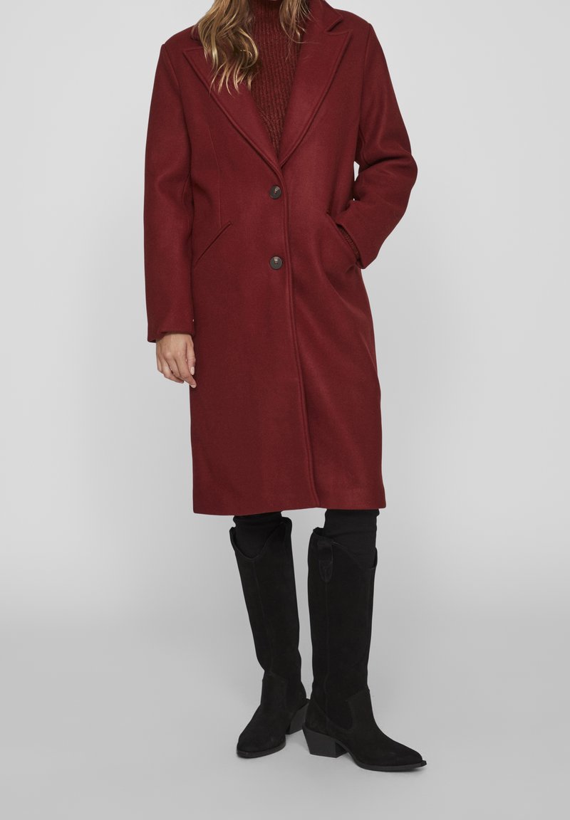 Manteau bordeaux en mélange de laine avec une fermeture à un bouton, des revers crantés et deux poches latérales. Associé à des bottes hautes en daim noir jusqu'aux genoux.