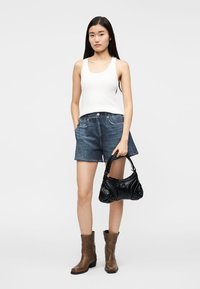 Pantaloncini di jeans blu, canottiera bianca a coste, stivaletti marroni alla caviglia, tiene una borsa a mano in pelle nera con design curvo e dettagli testurizzati.