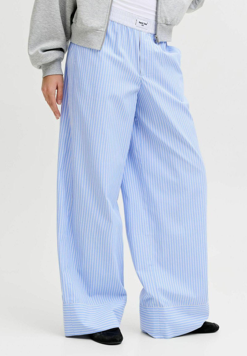 Personne portant un pantalon large à fines rayures bleu clair et blanc, une veste grise zippée et des chaussures noires, debout avec une main dans la poche.