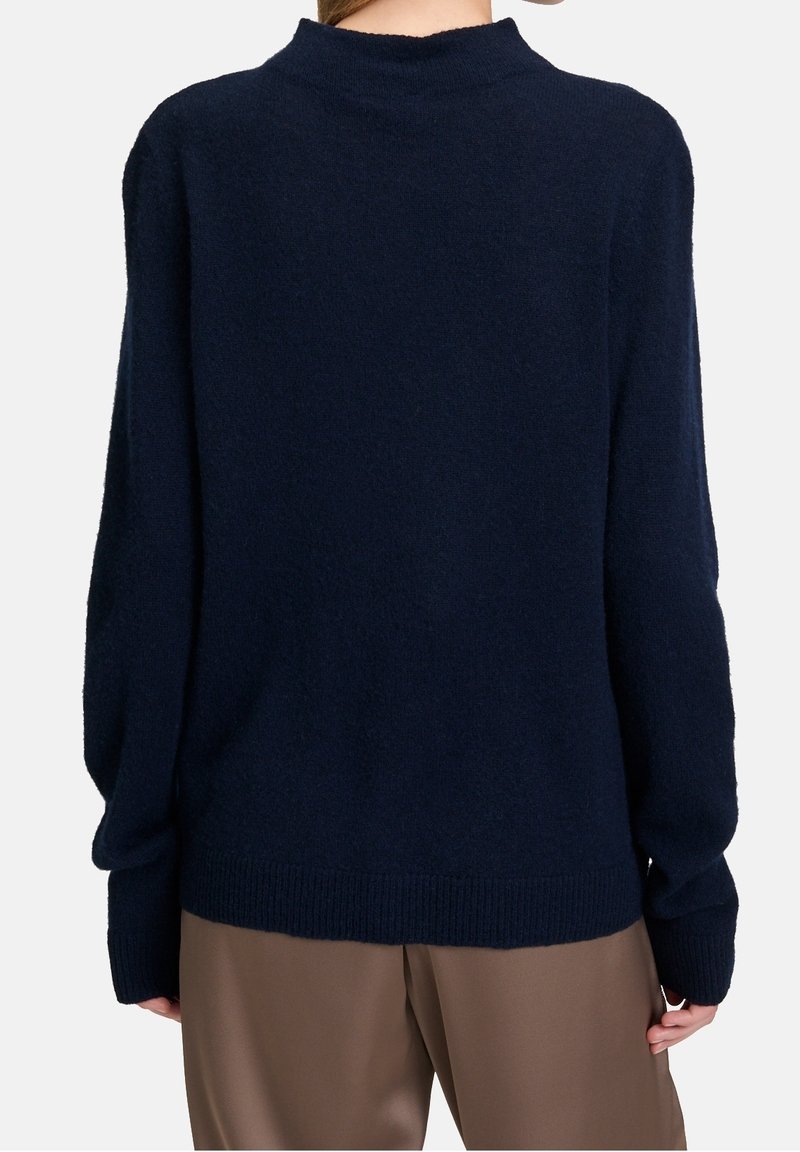 Personne portant un pull à manches longues bleu marine foncé et un pantalon ample marron, vue de dos sur un fond clair uni.