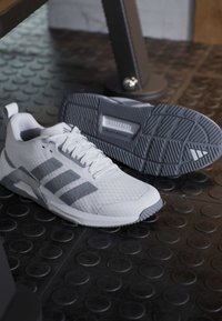 Chaussures de sport blanches et grises avec trois bandes, l’une vue de profil, l’autre montrant la semelle texturée sur un sol en caoutchouc noir.