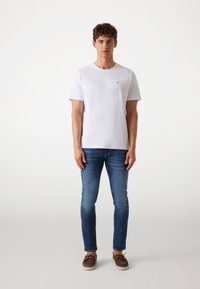 Witte katoenen T-shirt met korte mouwen, voorzien van een klein driehoekig embleem op de borst. Draagt met blauwe jeans en bruine schoenen.