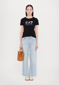 Zwarte t-shirt met "EA7 Emporio Armani" logo, lichtblauwe wijde jeans, en een geweven tan handtas, gecombineerd met crèmekleurige sneakers.