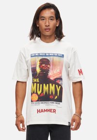 T-shirt en coton blanc featuring un imprimé de poster de film rétro pour "La Momie", avec des couleurs vives, du texte graphique et un logo sur la manche.