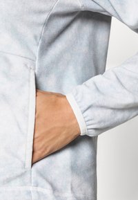 Svetlo modra, mehka flagra z žepom. Manšete so elastične z belim robom. Opazna je subtilna tie-dye tekstura.