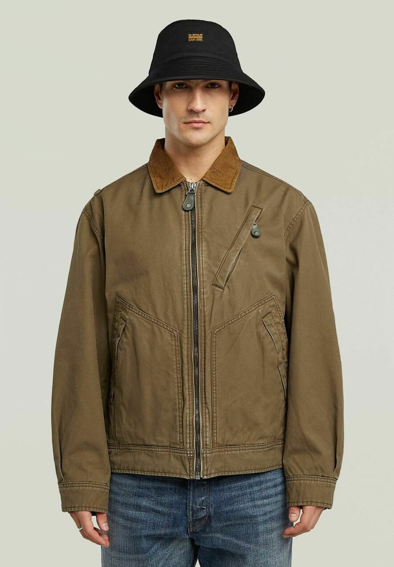 Veste en toile marron avec un col en velours côtelé, fermeture éclair, poches avant et un subtil motif diagonale ; assortie à un chapeau seau noir.