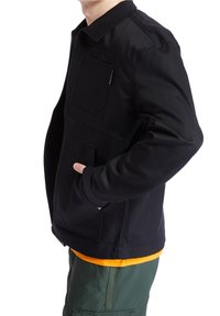 Černá bunda s předním zipem a dvěma náprsními kapsami, s klasickým límcem a hladkým povrchem, nošená přes oranžovou košili.