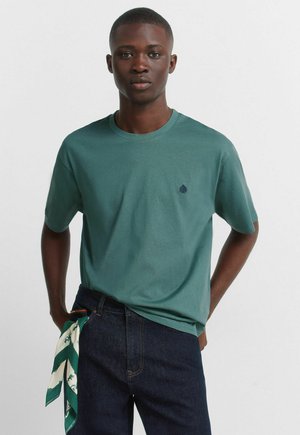 Jonge man staat met handen achter de rug, draagt een blauwgroen T-shirt en donkere spijkerbroek met een groen-wit patroon sjaal die uit zijn zak hangt.