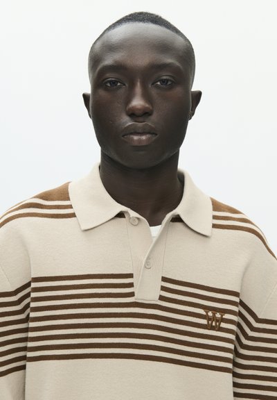 Polo rayé beige et marron en tissu doux, avec un col, deux boutons et un petit logo sur le côté gauche de la poitrine.