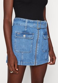 Lichtblauwe denim mini-jurk met een rits aan de voorkant, twee voorzakken en een riem. Voorzien van zilverkleurige hardware en een licht versleten textuur.