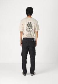 Beige t-shirt med sort blomsterkrukke-graphic på ryggen, parret med sorte cargo-bukser med sidelommer og en slim fit.