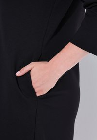 Czarna sukienka z boczną kieszenią, zbliżenie na rękę spoczywającą wewnątrz. Gładki materiał z dopasowanym krojem i długimi rękawami.