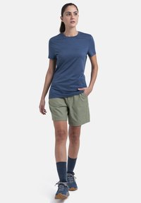 Blå kortærmet skjorte parret med grønne shorts, der har en afslappet pasform. Fodtøjet inkluderer grå vandresko og blå sokker, som fremhæver udendørsstilen.
