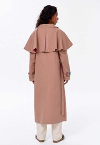 Kobieta z kręconymi włosami, ubrana w długi beżowy płaszcz z kapturem, beżowe spodnie i kapcie w panterkę, stojąca tyłem.