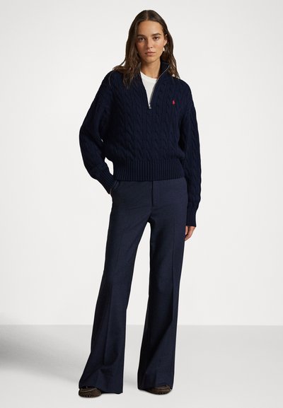 Γυναίκα που φοράει μπλε σκούρο πλεκτό πουλόβερ με φερμουάρ μέχρι τη μέση, λευκό υποκάμισο, φαρδιά μπλε σκούρα παντελόνια και καφέ παπούτσια, στέκεται μπροστά σε απλό φόντο.