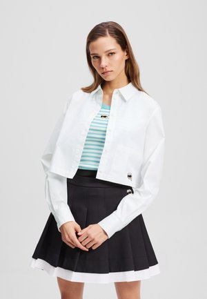 Cropped white button-up shirt over a blue and white striped tank top, paired with a black pleated skirt featuring white accents and patches.