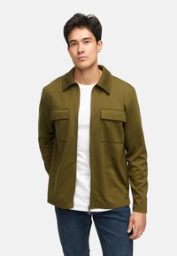 Giacca con zip verde oliva con colletto, due tasche frontali e una texture liscia, indossata sopra una camicia bianca e abbinata a jeans blu.