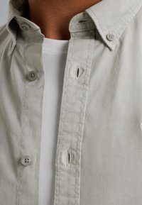 Camisa de botón gris claro con cuello, que presenta dos botones y una línea cosida por delante, superpuesta a una camisa blanca.