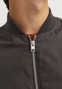 Black bomber jacket featuring a zip front, ribbed collar, and smooth texture. A contrasting orange garment is visible beneath the jacket.
