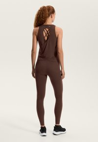 Rjav športni top z vzorcem križnih trakov na hrbtu, v kombinaciji s tesno prilegajočimi rjavimi legicami in črnimi športnimi čevlji. Gladek tekstura.