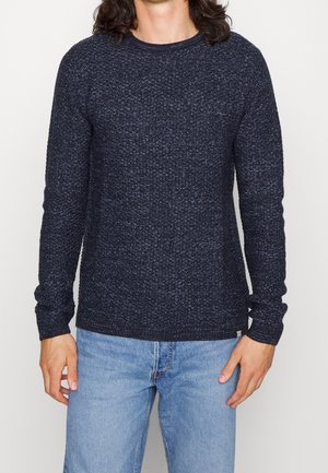 Uomo che indossa un maglione a maglia testurizzato blu scuro e jeans azzurri, in piedi contro uno sfondo bianco uniforme.