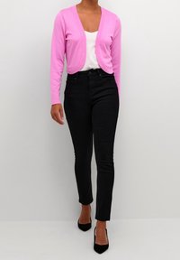 Růžová pletená cardigan s krátkým střihem, kulatým výstřihem a dlouhými rukávy, doplněná o černé skinny džíny a černé vysoké podpatky.
