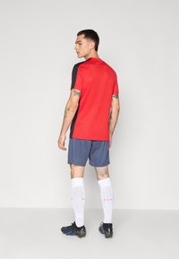 T-shirt de sport rouge avec des accents noirs, associé à un short bleu, des chaussettes blanches et des crampons noirs. Présente des tatouages sur les bras.