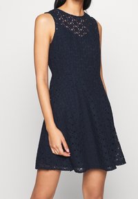 Ärmlös marinblå klänning i spetsmaterial, med en scalloped halsringning, strukturerat blommönster och en utsvängd kjol.