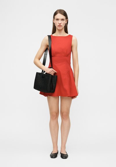 Γυναίκα που στέκεται φορώντας ένα κόκκινο αμάνικο φόρεμα, μαύρα επίπεδα παπούτσια και κρατώντας μια μαύρη υφασμάτινη τσάντα στον ώμο της, μπροστά από απλό φόντο.