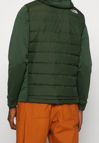 Vihreä puffertwesti tikatulla muotoilulla, kevyttä kangasta ja logo takana, kerroksittain tummanvihreän pitkähihasen paidan päällä.