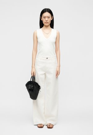 Femeie purtând un top alb fără mâneci cu decolteu în V și pantaloni albi largi, ținând o geantă neagră cu franjuri și purtând sandale.