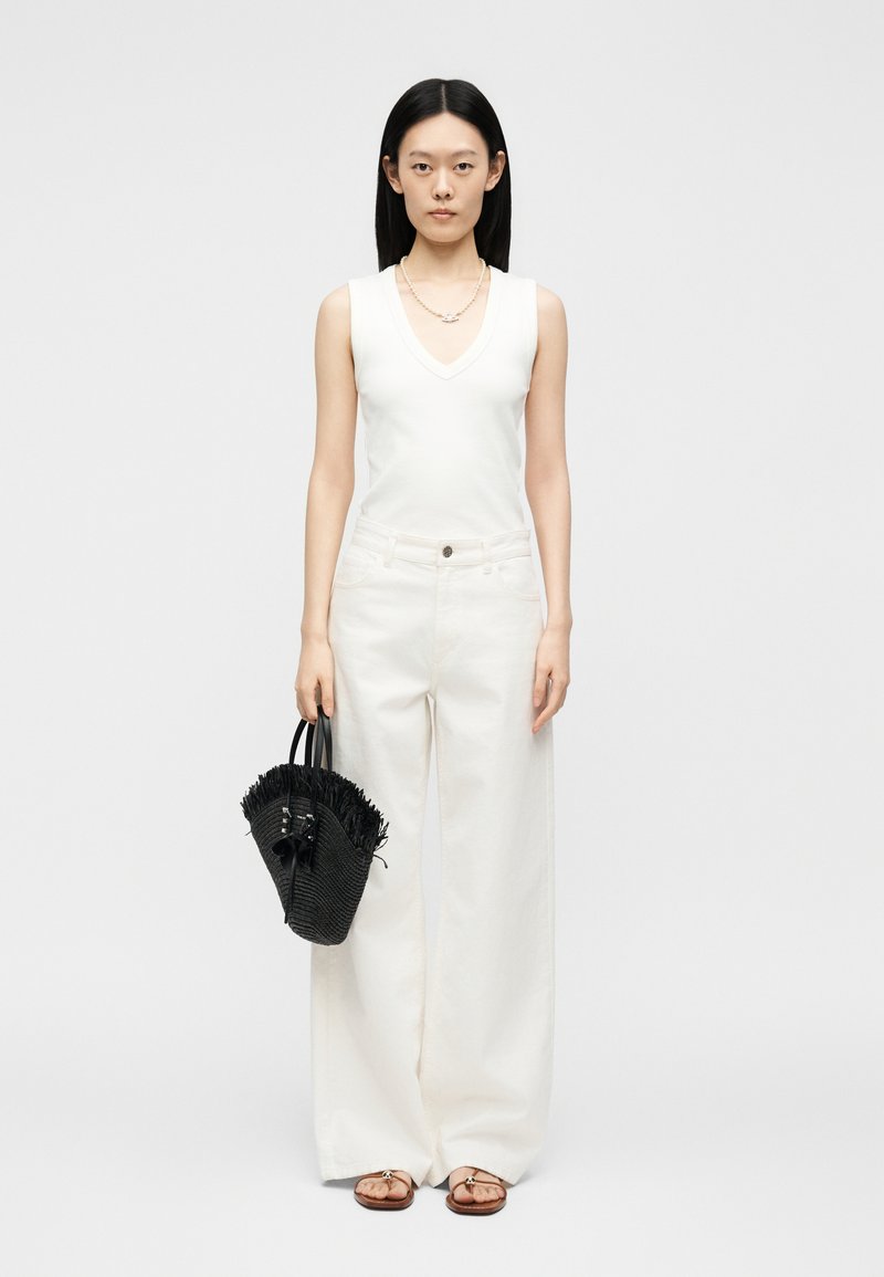 Γυναίκα που φοράει λευκό αμάνικο τοπ με V-λαιμόκοψη και φαρδύ λευκό παντελόνι, κρατώντας μια μαύρη τσάντα με φουντίτσες και φοράει σανδάλια.