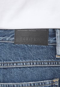Jeans de denim con un parche de cuero oscuro etiquetado "CLOSED" en la cintura. Presenta un denim azul descolorido y costuras en contraste.