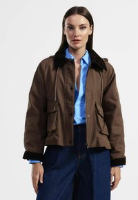 Giubbotto bomber marrone con colletto in velluto a coste, due tasche frontali, chiusura con zip e polsini a costine, indossato sopra una camicia blu e jeans scuri.
