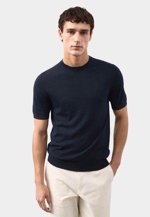Mand med kort krøllet hår iført en tætsiddende marineblå kortærmet sweater og lyse beige bukser, stående mod en ensfarvet baggrund.