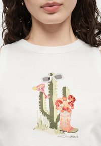 Κοντινό πλάνο ενός λευκού τοπ με γραφικό κάκτου που φοράει γυαλιά ηλίου και κρατάει ένα ροζ ποτήρι κοκτέιλ δίπλα σε μια λουλουδένια καουμπόικη μπότα.