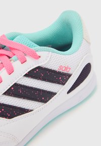Zapatilla deportiva blanca con cordones y detalles en rosa, con rayas negras salpicadas de rosa. Parte superior de malla texturizada y forro turquesa.
