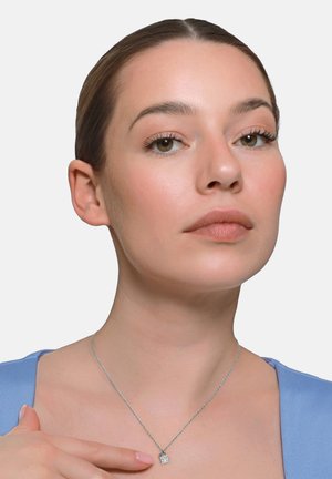 Frau mit zurückgebundenem Haar, die ein blaues Oberteil und eine silberne Halskette mit einem Vorhängeschloss-Anhänger trägt, berührt den Anhänger sanft.