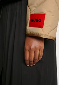 Brunet hud hånd afslappet ved siden, iført beige quiltet ærme med rødt mærke mærket "HUGO," mod sort plisseret stof baggrund.