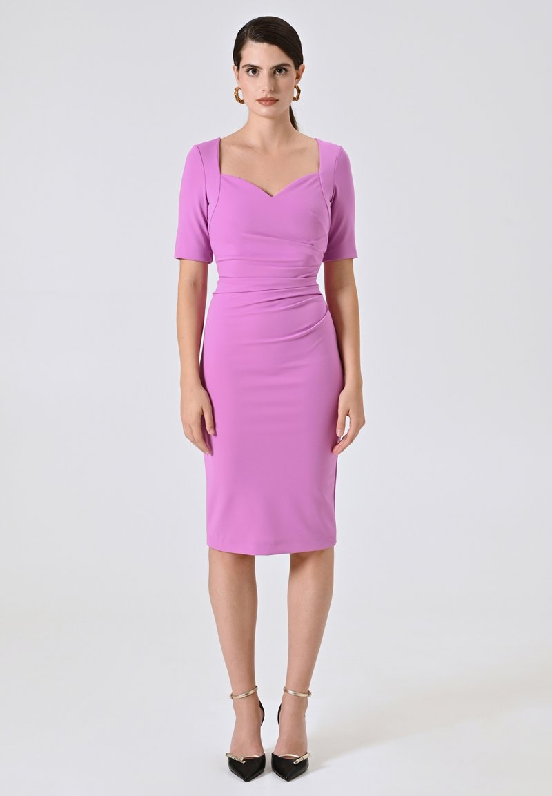 Aansluitende roze jurk met korte mouwen, vierkante halslijn en geplooide details in de taille. Draagt zwarte pumps met spitse neuzen.