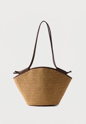 Sac fourre-tout en paille tressée avec garniture en cuir marron foncé et longues bandoulières réglables, forme trapèze sur fond blanc.