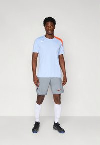 Camisola Nike de mangas curtas azul-claro com destaque laranja no ombro, combinada com calções cinzentos com logótipo vermelho, meias brancas e chuteiras pretas.