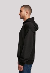 Sort hoodie med kængurulomme, lange ærmer og ribbede manchetter. Rent design med en blød stoftekstur, vist på en models sideprofil.